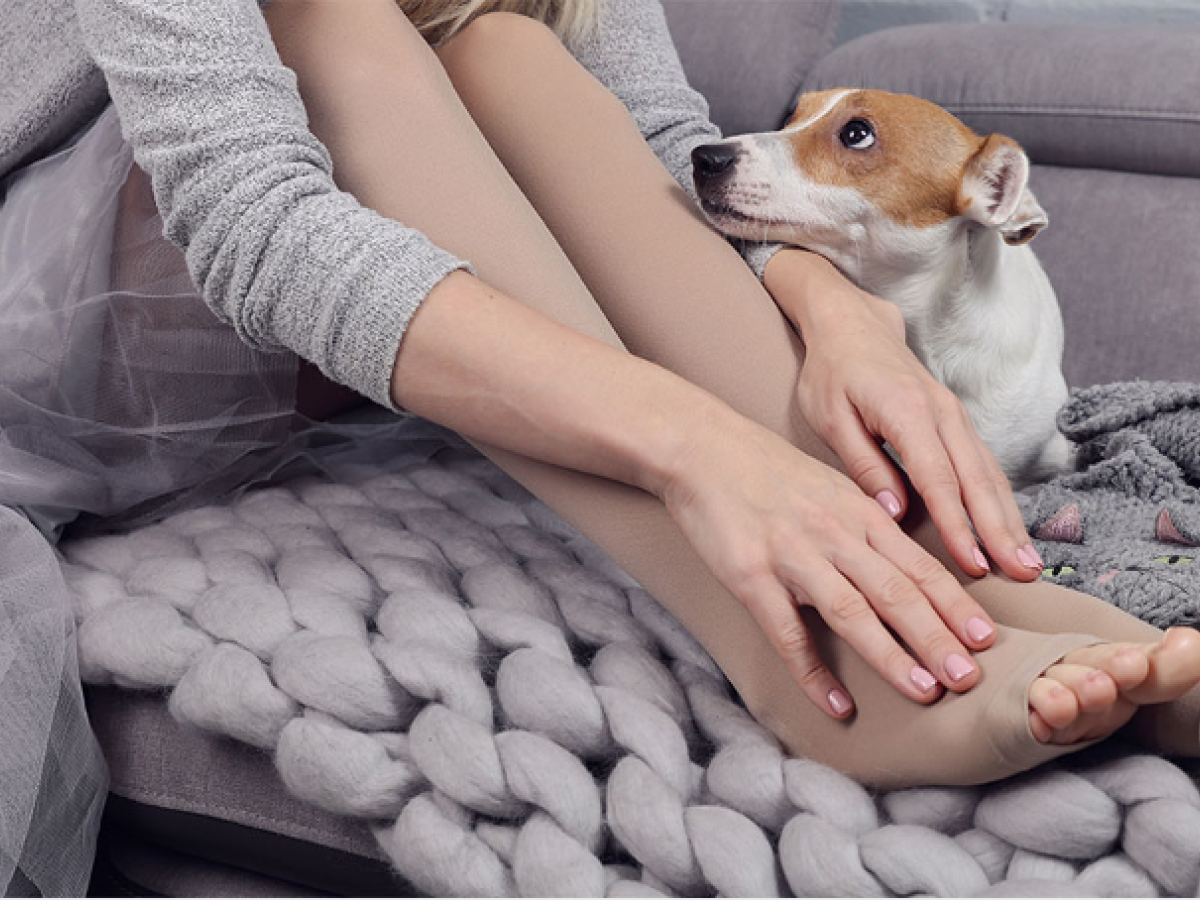 Eine Frau sitzt mit ihrem Hund auf einem Sofa und fasst sich an die geschwollenen Füße